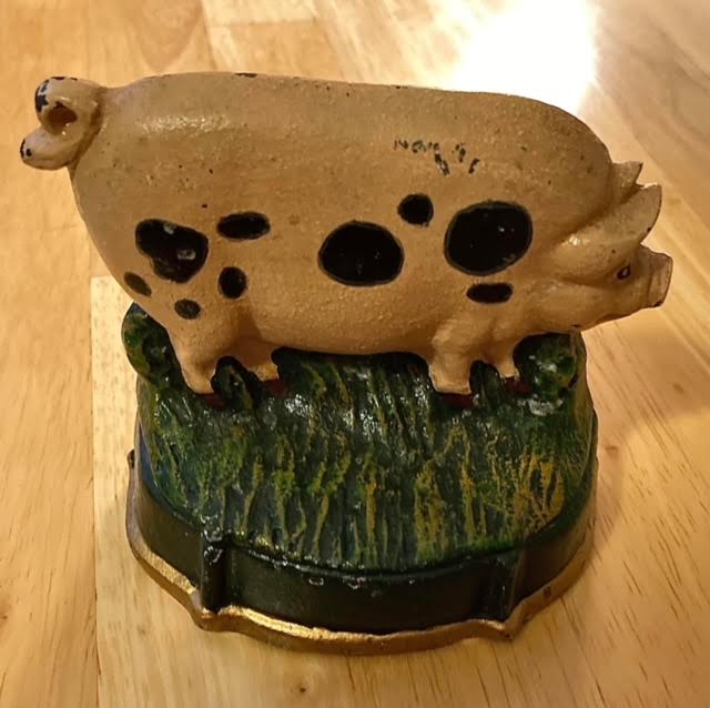 photograph of an iron pig doorstop. The pig is beige, with a large and small round black circles on its body. It's standing on a mound of green grass.